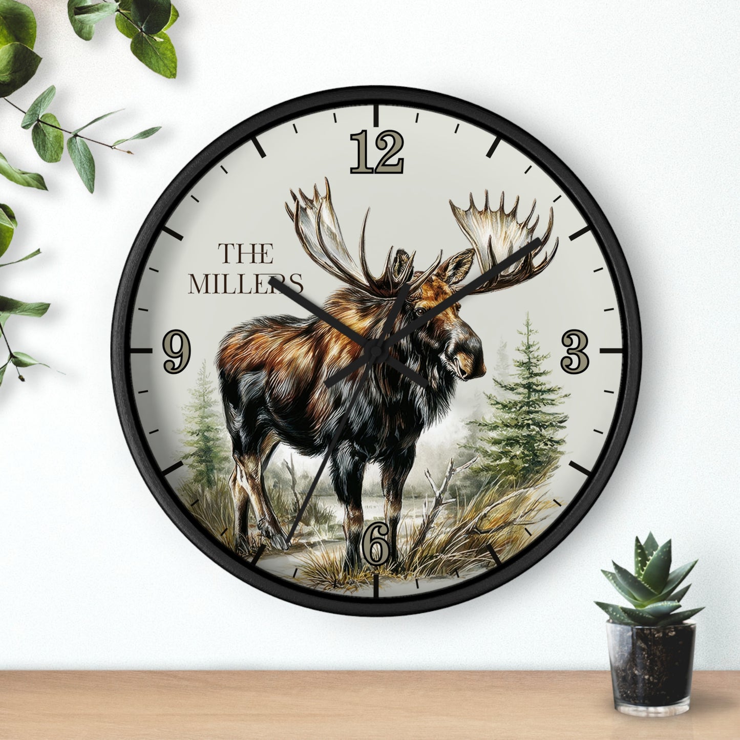A round wall clock with a black frame and a nature illustration of a moose on the dial, with the text 'THE MILLERS' and numerals 1 through 6. The clock is mounted on a white wall above a wooden surface, with decorative green leaves and a small potted succulent nearby.