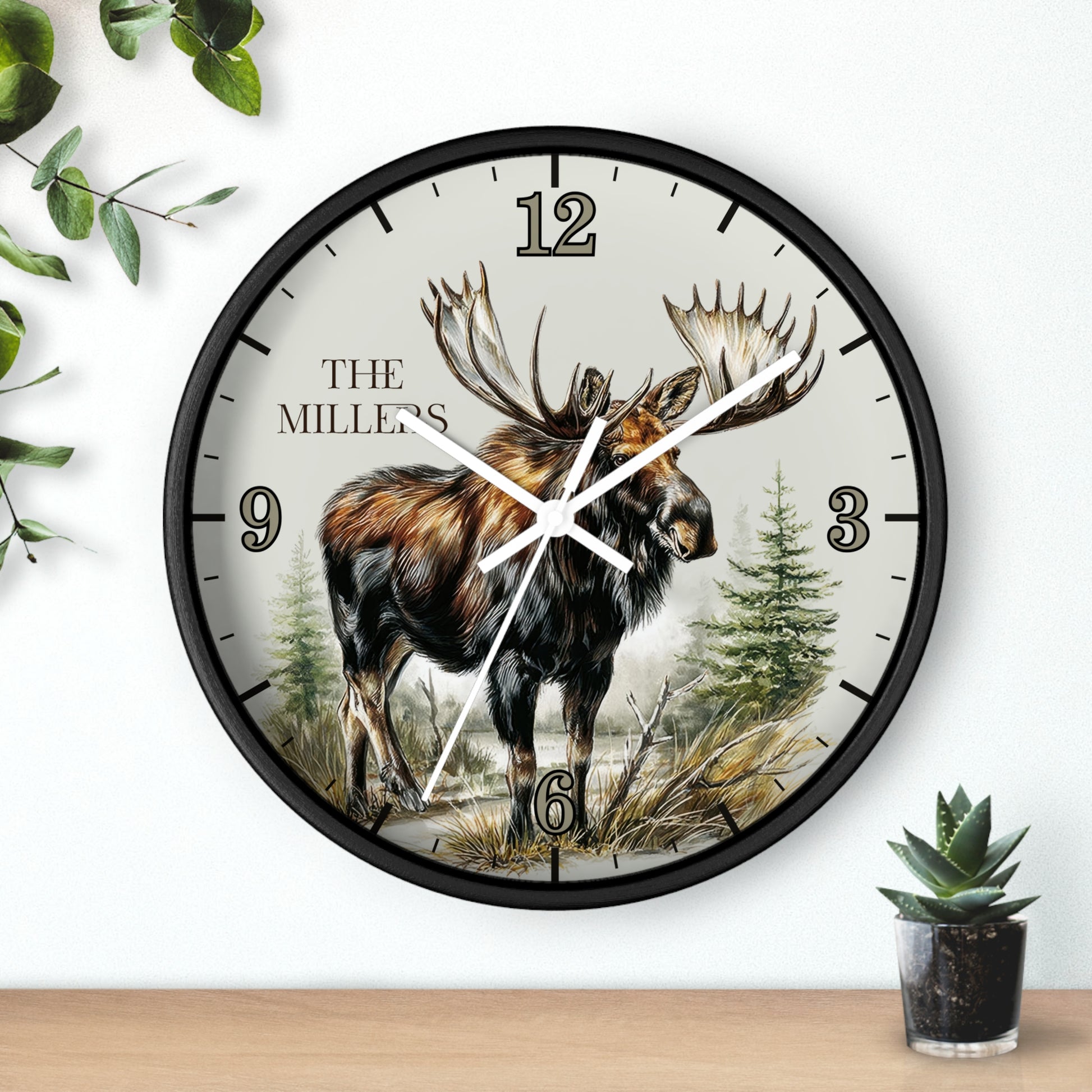A round wall clock with a black frame and a nature illustration of a moose on the dial, mounted on a white wall above a wooden surface with small green plants nearby.
