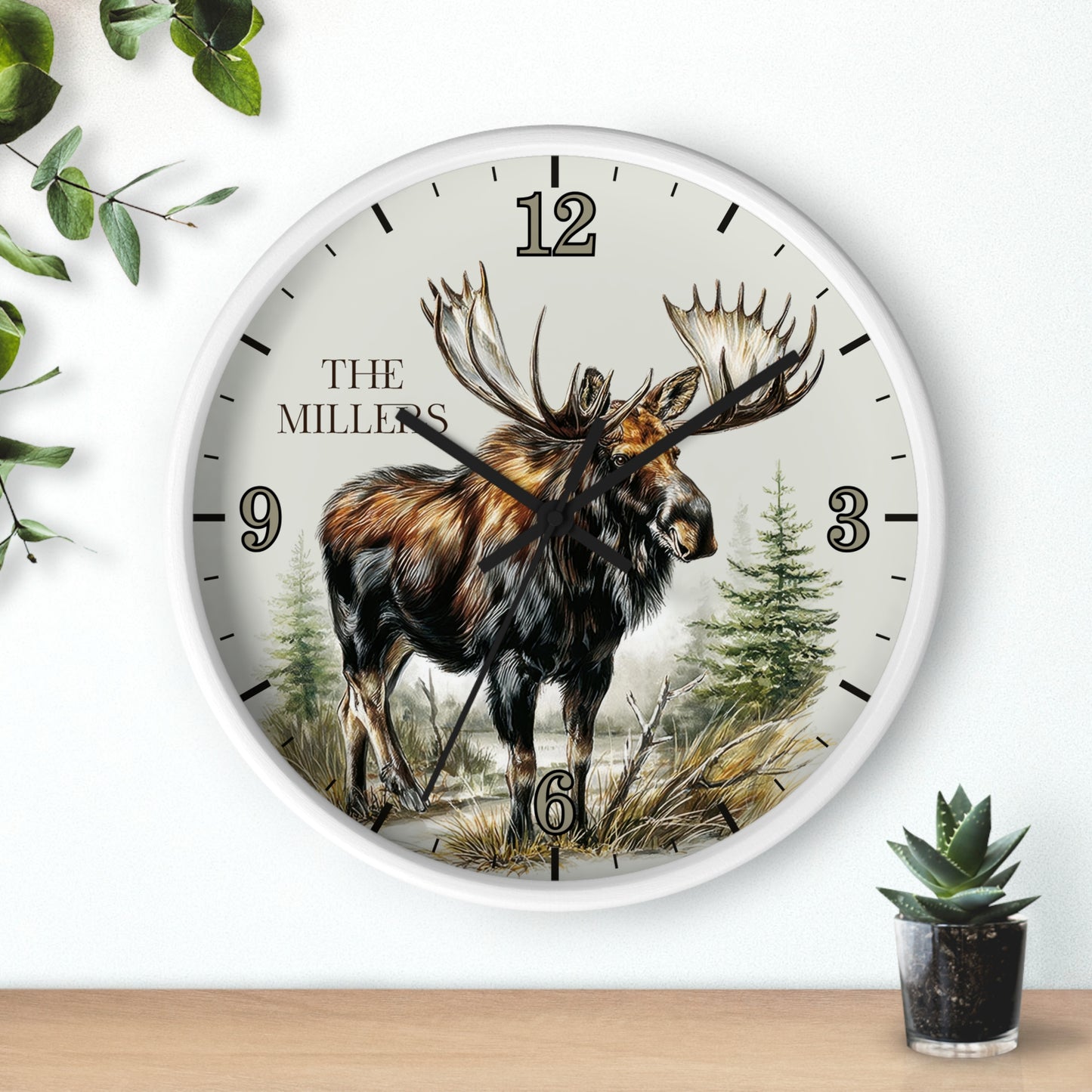 A round wall clock with a white frame and a detailed illustration of a moose on the dial, with the text 'THE MILLERS' and black Arabic numerals. The clock is mounted on a white wall above a light wood surface, with decorative green leaves and a small potted succulent nearby.