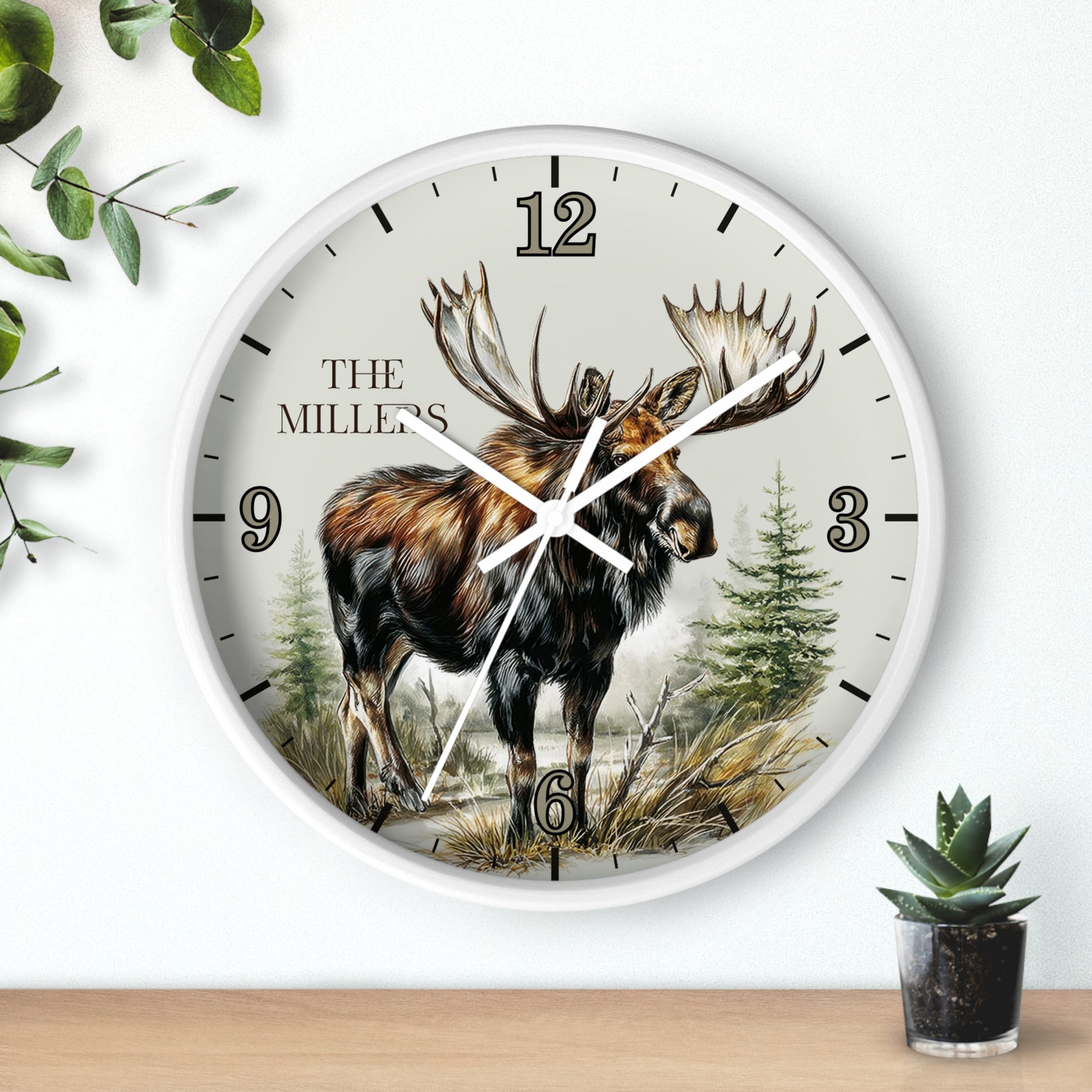 A round wall clock with a white frame and a detailed illustration of a moose on the face, with the text 'THE MILLERS' near the 12 o'clock position. The clock is mounted on a white wall above a light wood surface, with decorative green leaves and a small potted succulent nearby.