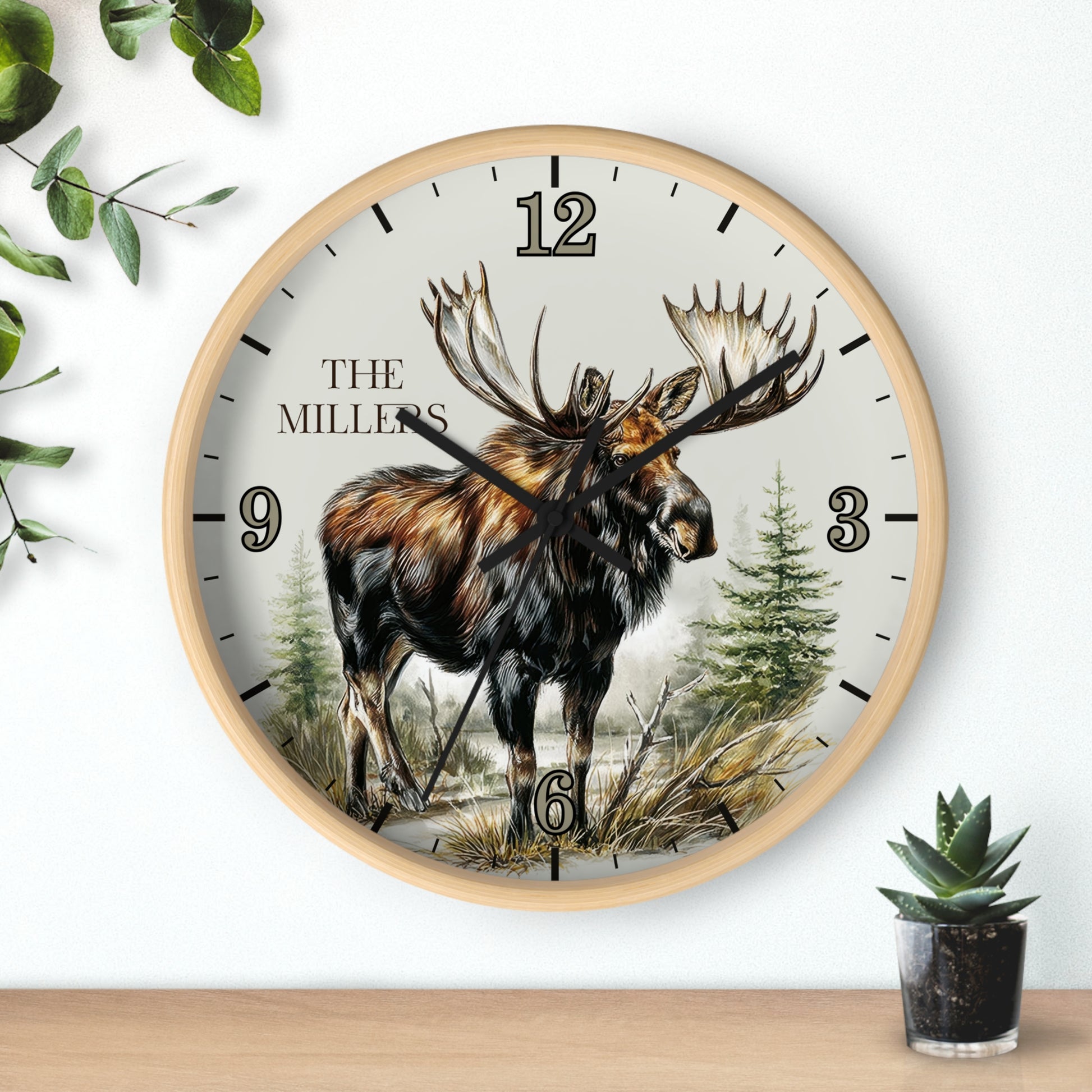 A round wall clock with a light wood frame and a nature illustration of a moose on the dial. The clock face is off-white with black Arabic numerals and the text 'THE MILLERS' near the 12.