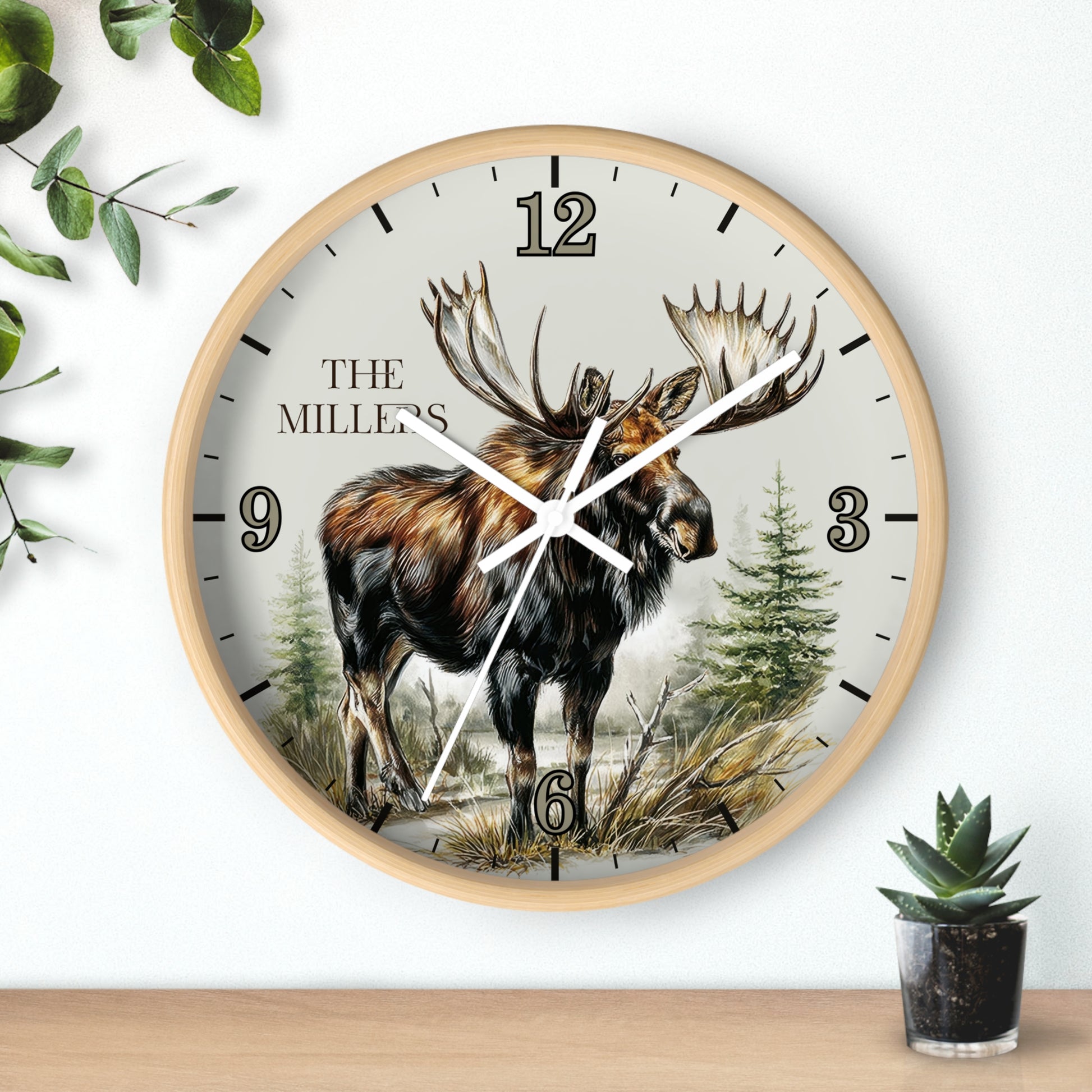 A round wall clock with a light wood frame and a nature illustration of a moose on the face, mounted on a white wall above a wooden surface with small green plants.