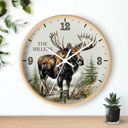 A round wall clock with a light wood frame and a nature illustration of a moose on the face, mounted on a white wall above a wooden surface with small green plants.
