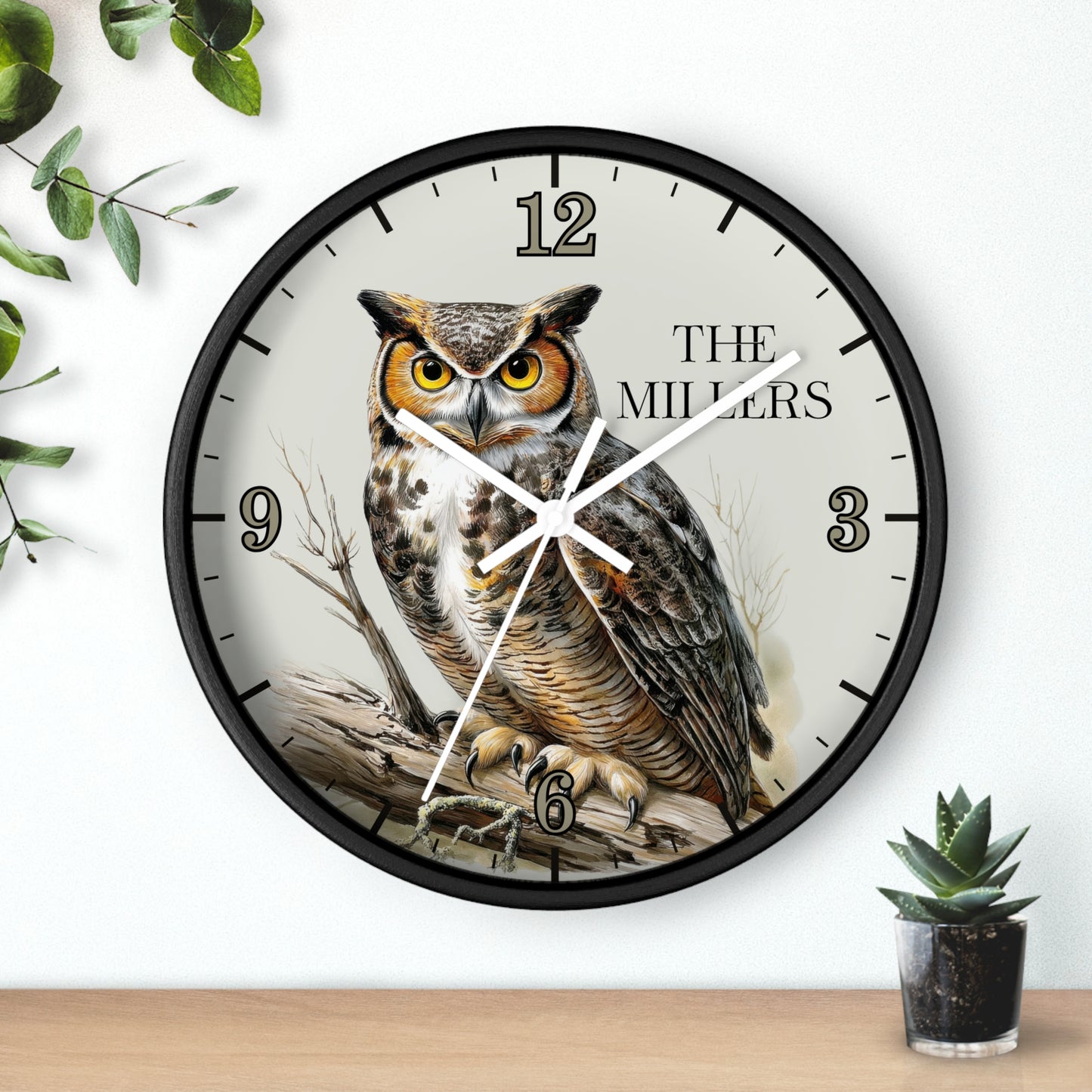 A round wall clock with a black frame and a printed owl illustration on the face, with the text 'THE MILLERS' and black Arabic numerals. The clock is mounted on a white wall above a light wood surface, with decorative green leaves and a small potted succulent nearby.