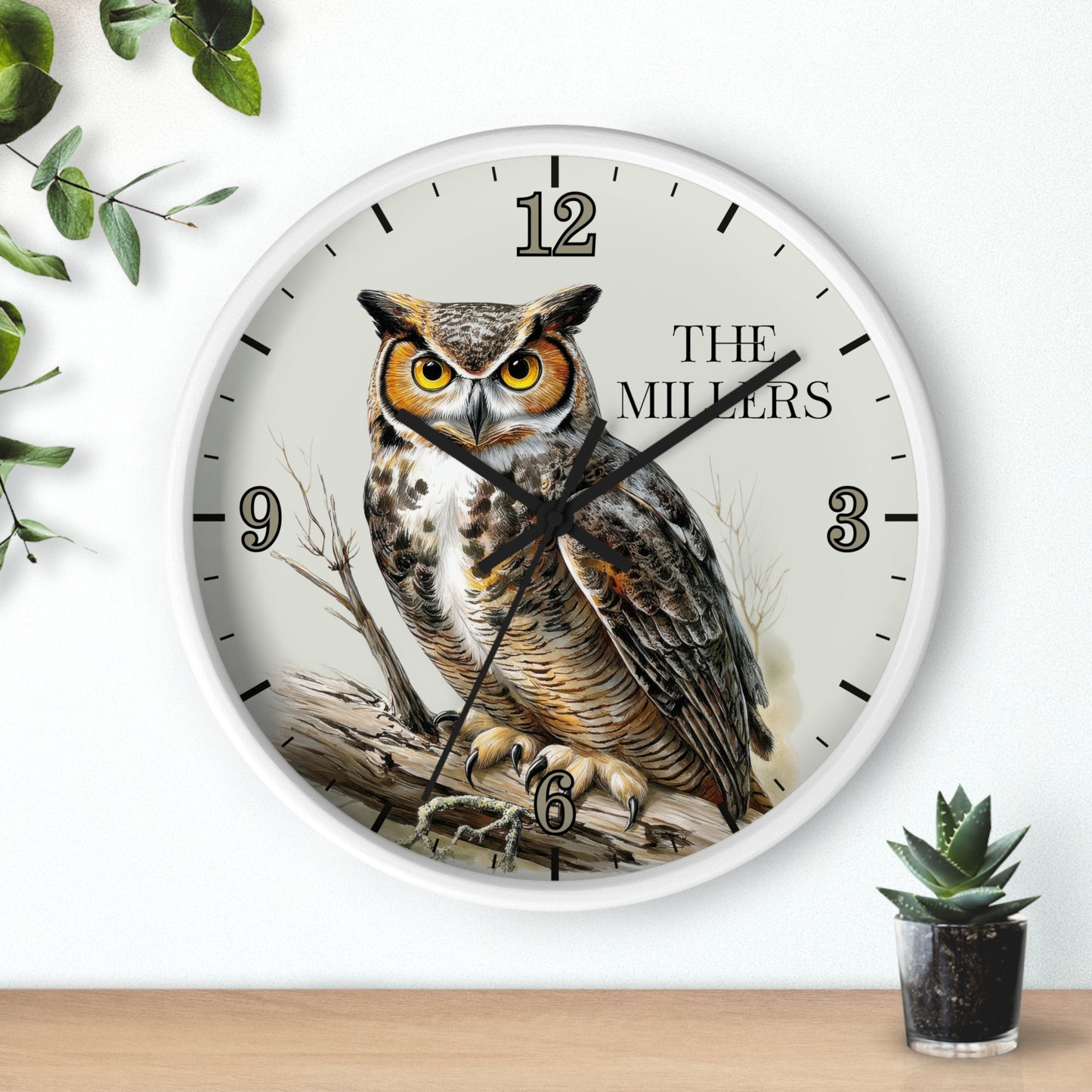 A round wall clock with a white frame and a detailed owl illustration on the face, with the text 'THE MILLERS' and clock numbers. The clock is mounted on a white wall above a light wood surface, with decorative green leaves and a small potted succulent nearby.