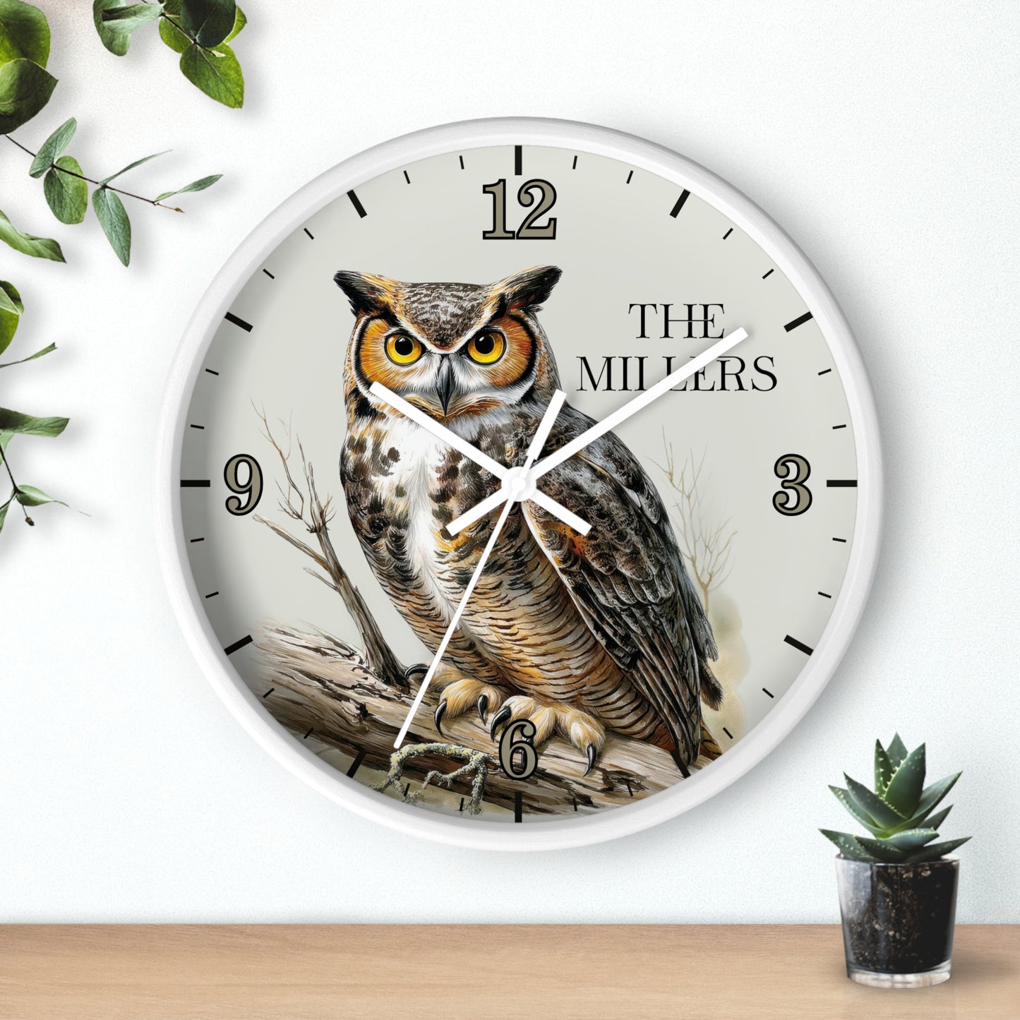 A round wall clock with a white frame and a printed owl illustration on the face, mounted on a white wall above a wooden surface with small green plants nearby.