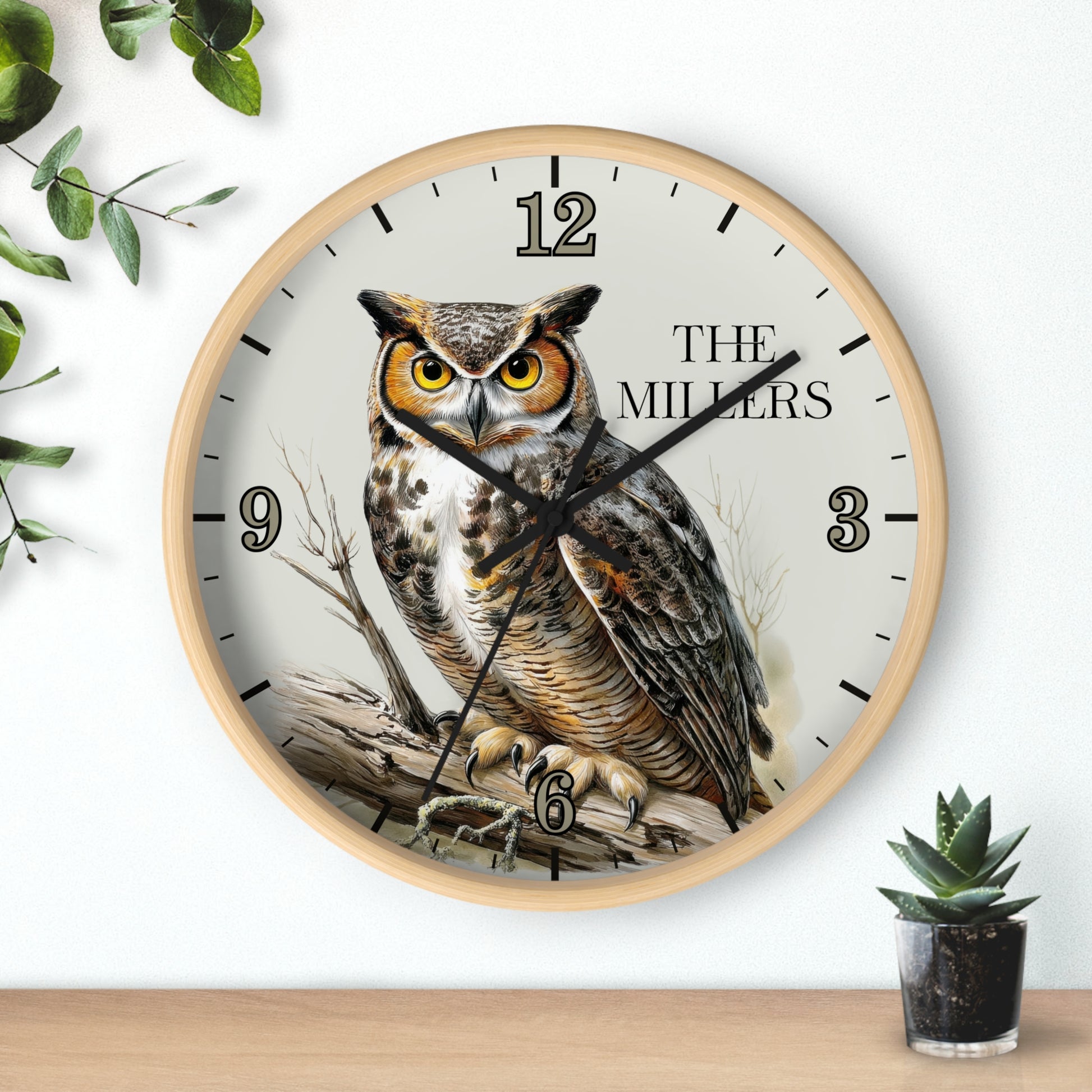 A round wall clock with a light wood frame and a printed owl illustration on the face, with black numerals and hands. The clock is mounted on a white wall above a wooden surface, with decorative green leaves and a small potted succulent nearby.