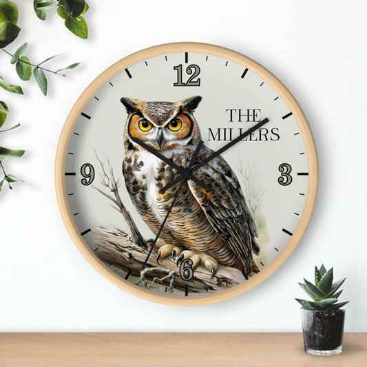 A round wall clock with a light wood frame and a printed owl illustration on the face, with black numerals and hands. The clock is mounted on a white wall above a wooden surface, with decorative green leaves and a small potted succulent nearby.