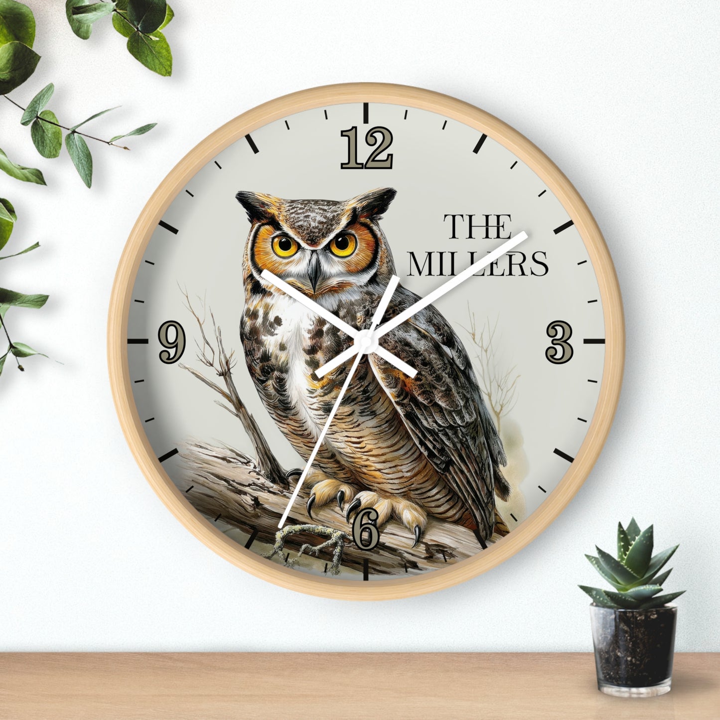 A round wall clock with a light wood frame and a printed owl illustration on the face, with black Arabic numerals and the text 'THE MILLER' on a white background. The clock is shown hanging on a white wall above a wooden surface, with decorative green leaves and a small potted succulent nearby.