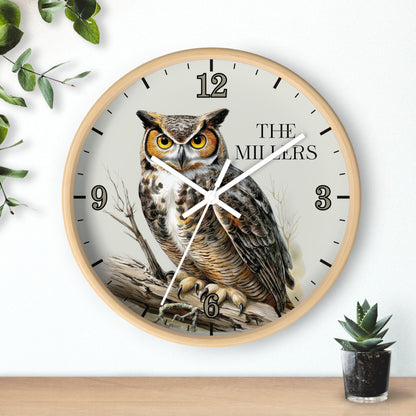 A round wall clock with a light wood frame and a printed owl illustration on the face, with black Arabic numerals and the text 'THE MILLER' on a white background. The clock is shown hanging on a white wall above a wooden surface, with decorative green leaves and a small potted succulent nearby.