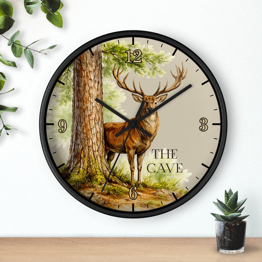 A round wall clock with a black frame and a printed forest scene featuring a stag standing beside a tree. The clock face has black numerals and hands, with the words 'THE CAVE' printed near the lower right.