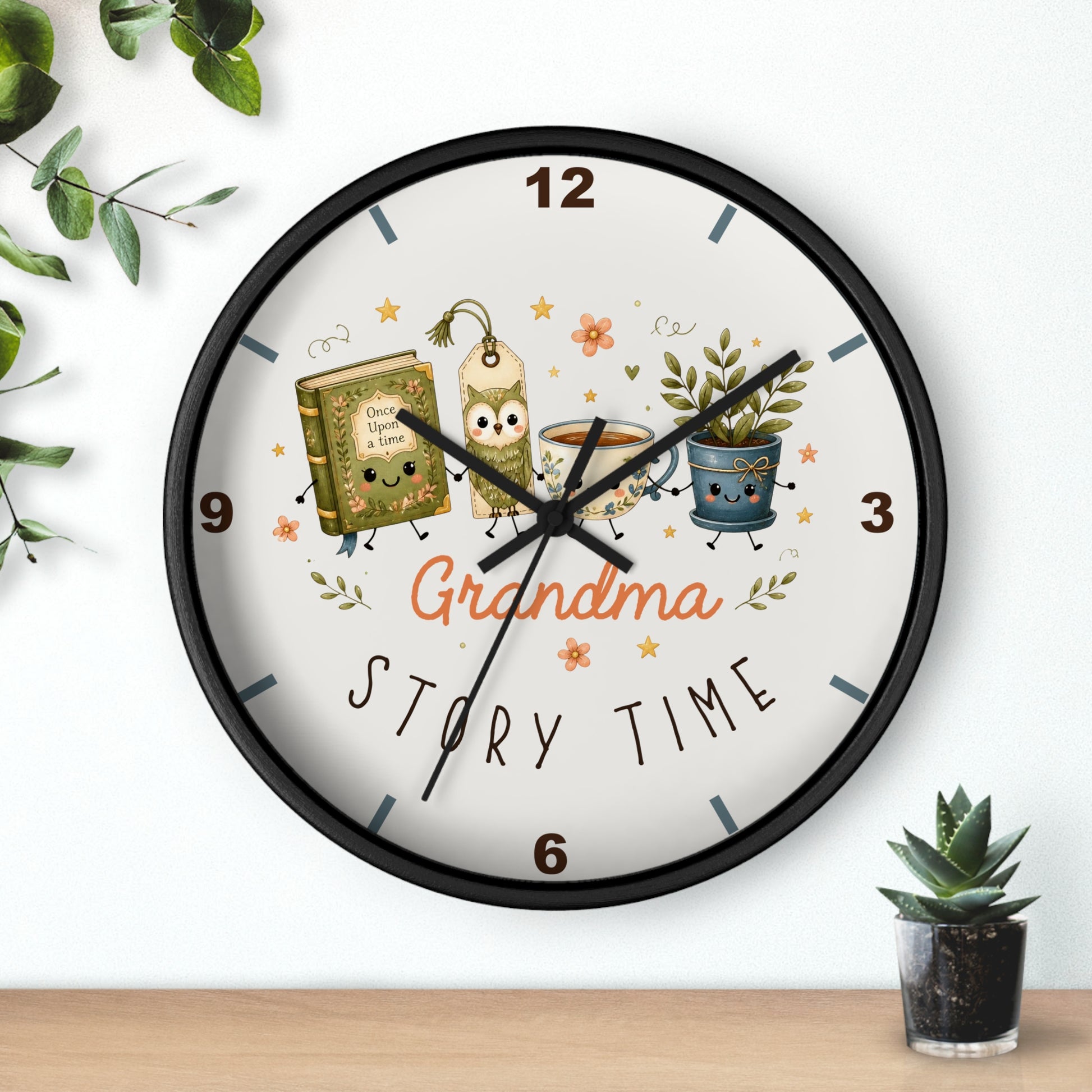 A round wall clock with a black frame and a white face, featuring a whimsical 'Grandma Story Time' illustration with cartoon characters, a book, a mug, and potted plants. The clock is shown hanging on a white wall above a wooden shelf with small green plants.