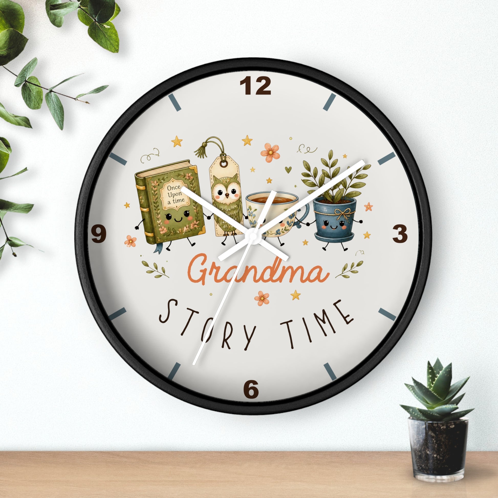 A round wall clock with a black frame and a white face, decorated with cartoon illustrations of a book, a mug, and a potted plant, along with the words 'Grandma Story Time'. The clock is shown hanging on a white wall above a wooden shelf with small green plants.