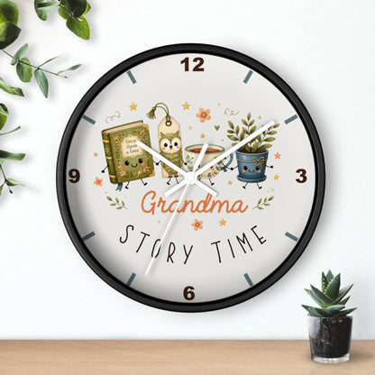 A round wall clock with a black frame and a white face, decorated with cartoon illustrations of a book, a mug, and a potted plant, along with the words 'Grandma Story Time'. The clock is shown hanging on a white wall above a wooden shelf with small green plants.