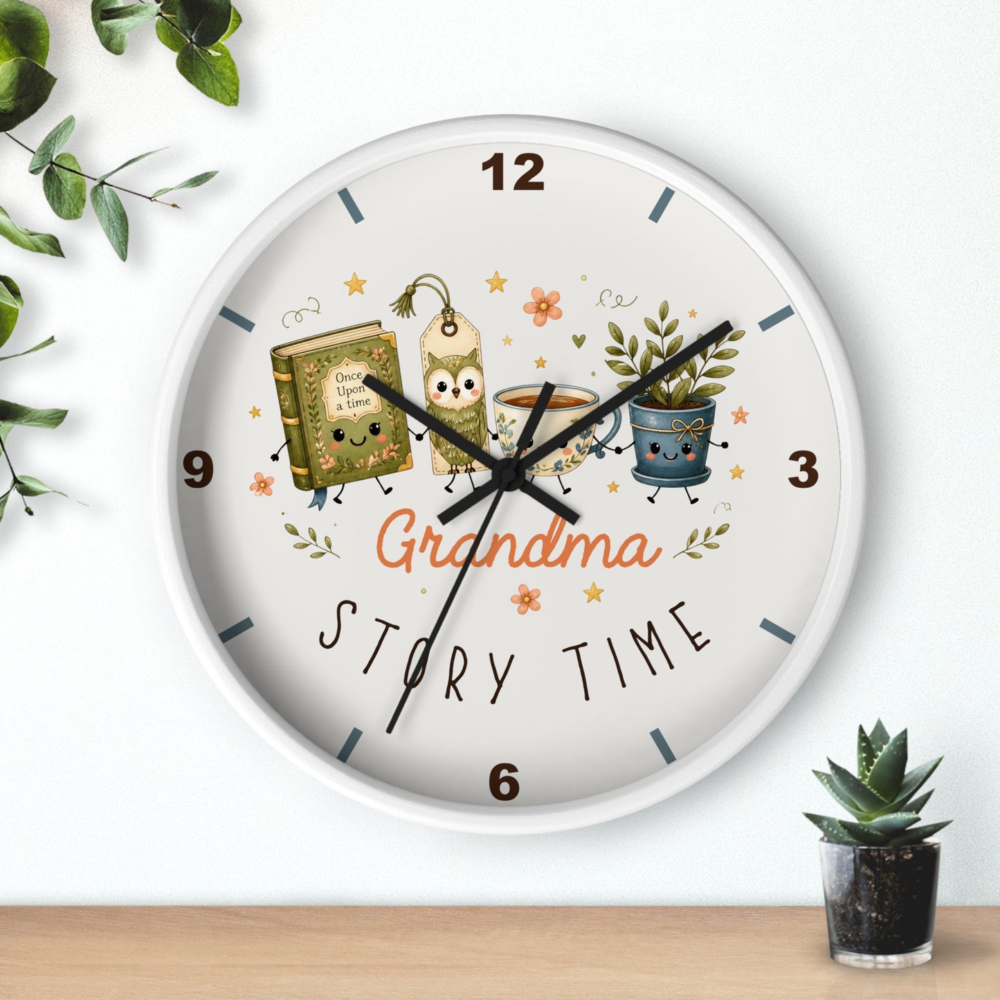A round wall clock with a white face and silver rim, featuring a whimsical 'Grandma Story Time' illustration with a book, mug, and potted plant. The clock is mounted on a white wall above a wooden shelf with small green plants.