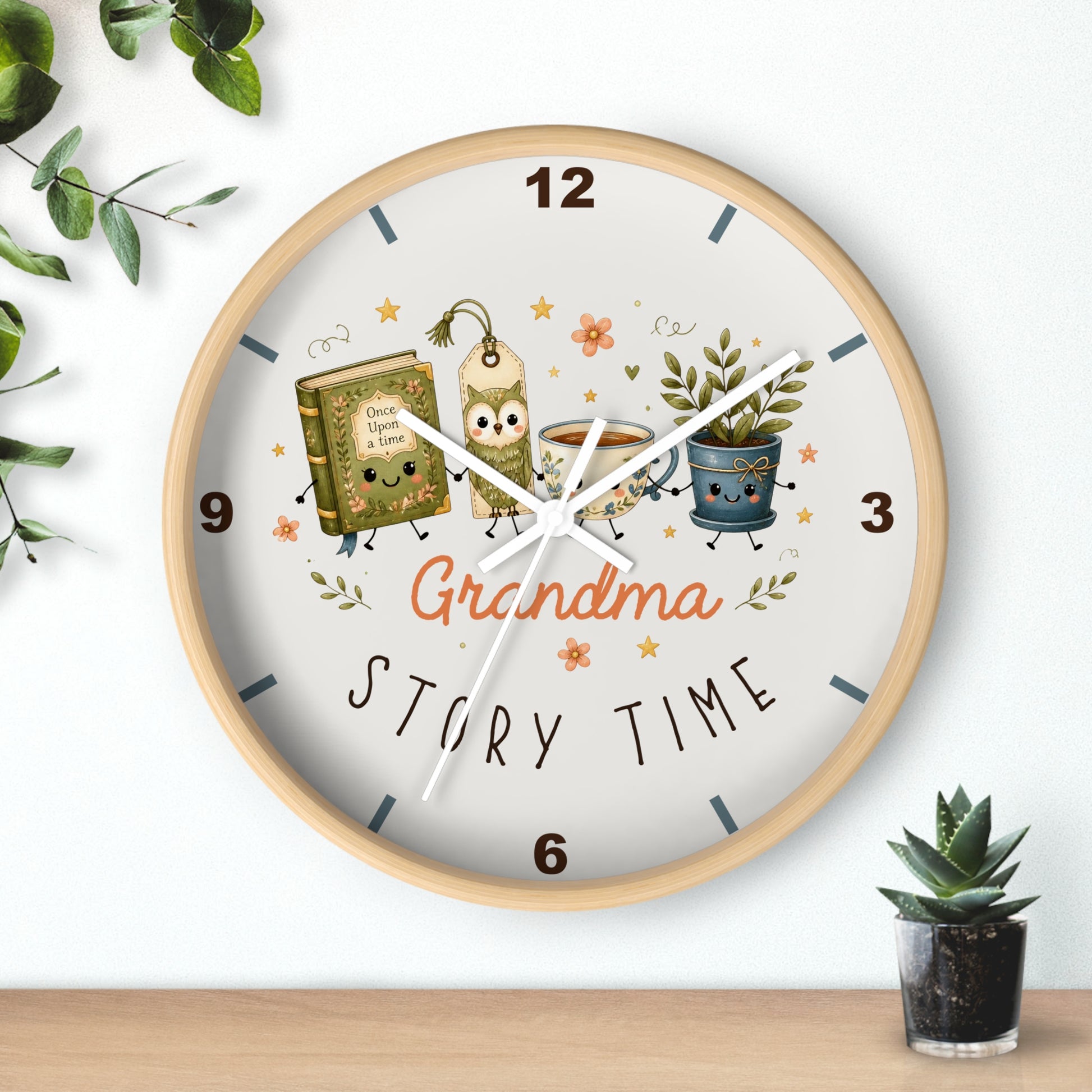 A round wall clock with a light wood frame and a white face, decorated with cartoon illustrations of a book, a coffee mug, and a potted plant, with the words 'Grandma Story Time' in the center. The clock is mounted on a white wall above a wooden shelf, with decorative green leaves and a small succulent plant nearby.