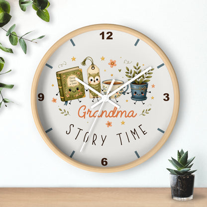 A round wall clock with a light wood frame and a white face, decorated with cartoon illustrations of a book, a coffee mug, and a potted plant, with the words 'Grandma Story Time' in the center. The clock is mounted on a white wall above a wooden shelf, with decorative green leaves and a small succulent plant nearby.