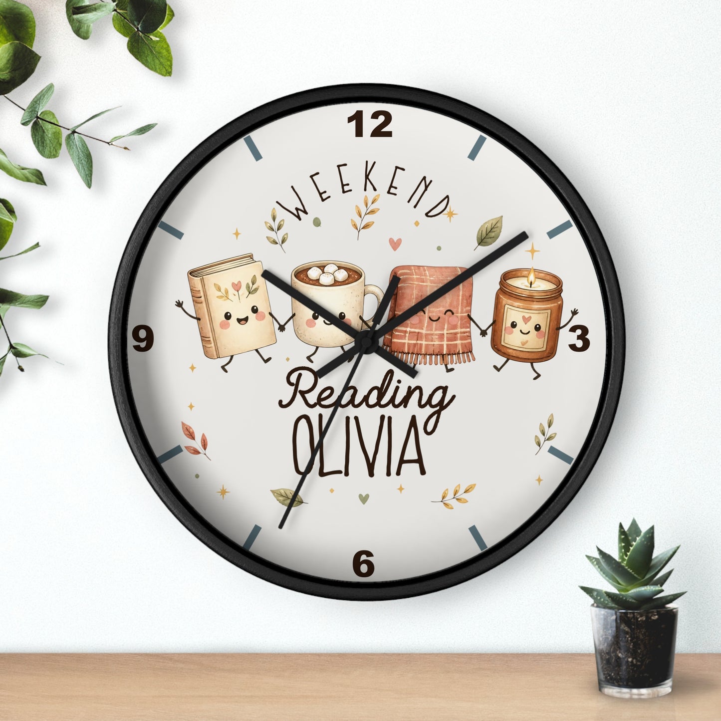 A round wall clock with a black frame and a white face, decorated with cute illustrations of books, a mug, a blanket, and a candle, along with the text 'WEEKEND Reading OLIVIA'. The clock is shown hanging on a white wall above a wooden shelf with small decorative plants.
