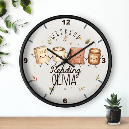 A round wall clock with a black frame and a white face, decorated with cute illustrations of books, a mug, a blanket, and a candle, along with the text 'WEEKEND Reading OLIVIA'. The clock is shown hanging on a white wall above a wooden shelf with small decorative plants.