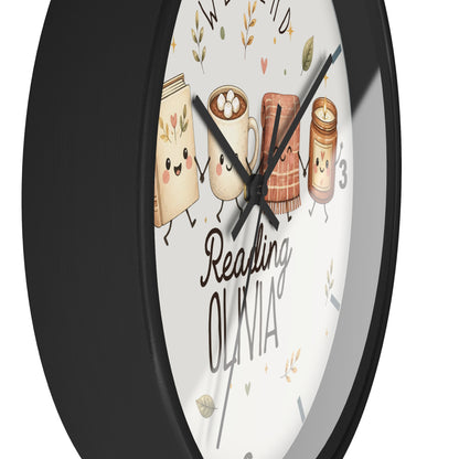 A close-up of a round wall clock with a black frame and a white face. The clock face features cute illustrations of a book, a mug, a scarf, and a candle, along with the text 'Reading OLIVIA' in black script and block lettering.