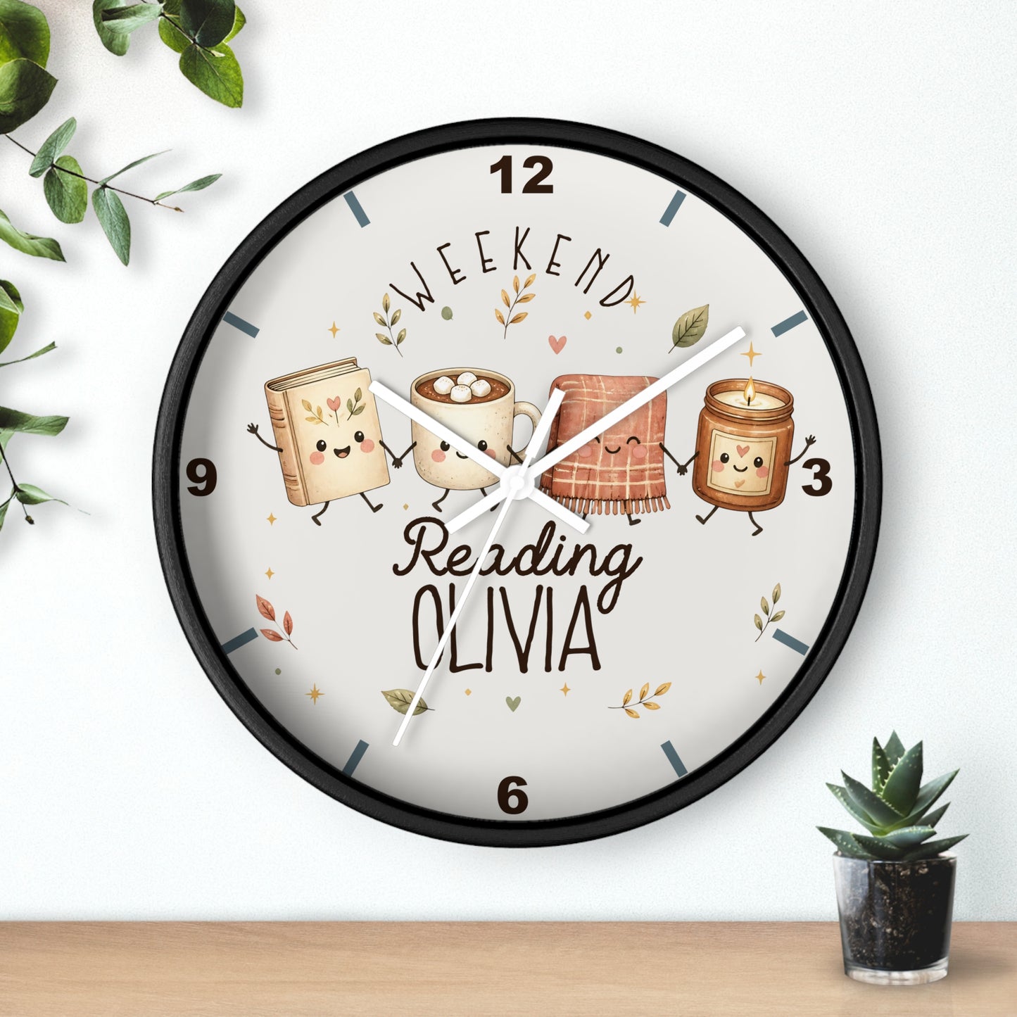 A round wall clock with a black frame and a white face, featuring a whimsical illustration of four marshmallows as books with the text 'WEEKEND Reading OLIVIA'. The clock is mounted on a white wall above a wooden shelf, styled with small green plants.