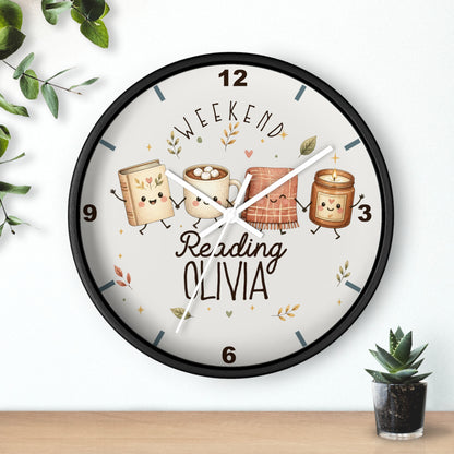 A round wall clock with a black frame and a white face, featuring a whimsical illustration of four marshmallows as books with the text 'WEEKEND Reading OLIVIA'. The clock is mounted on a white wall above a wooden shelf, styled with small green plants.