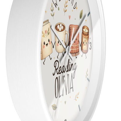 A round white wall clock with a whimsical illustration of reading-themed characters on the face, including a book, a mug with marshmallows, a slice of toast, and a candle jar. The words 'Reading OLIVIA' are printed on the dial.