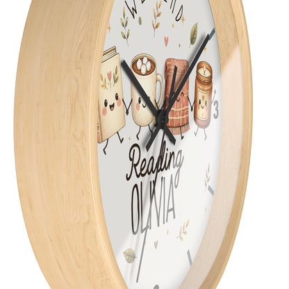 A round wall clock with a light natural wood frame and a white face featuring cute illustrated books, a mug, a scarf, and a candle, plus the text 'Reading OLivia'.