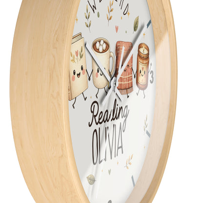A round wall clock with a light natural wood frame and a white face featuring cute illustrated books, a mug, a loaf of bread, and a candle, plus the text 'Reading OLIVIA'.