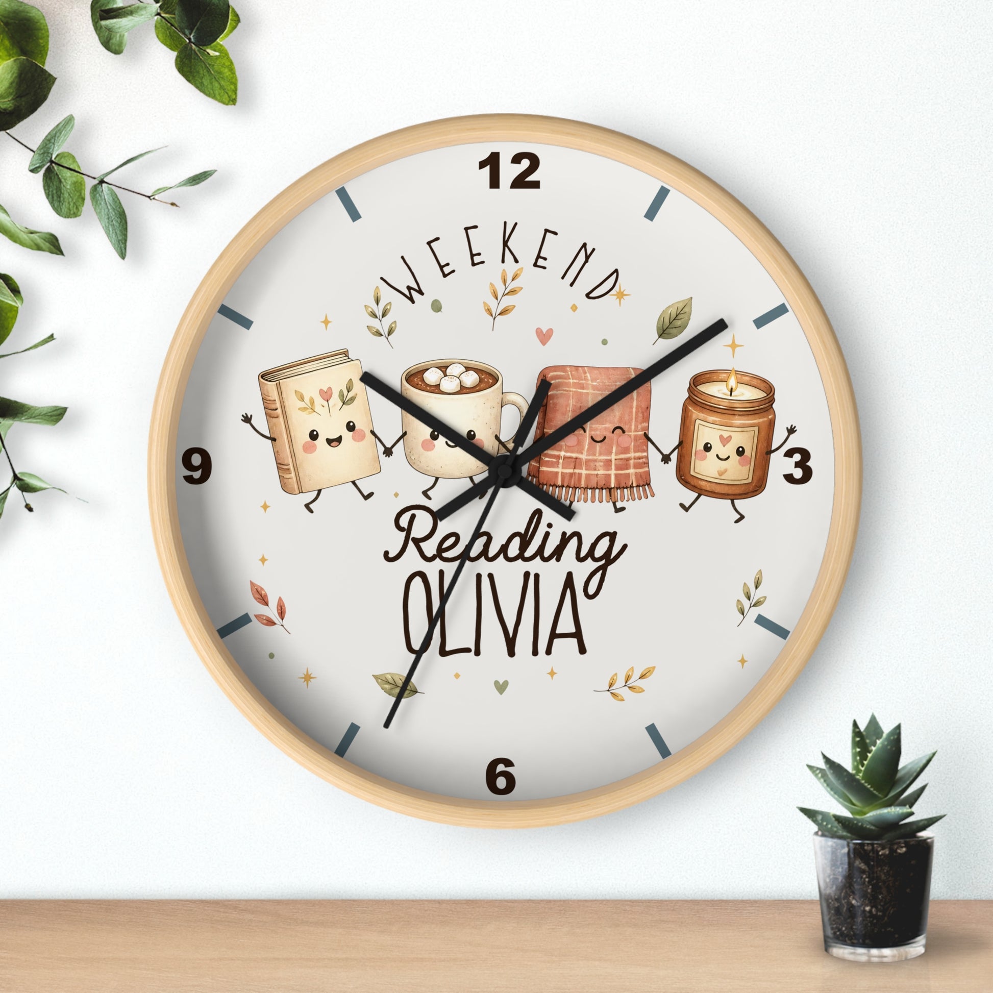 A round wall clock with a light wood frame and a white face, featuring a cute illustration of four books and a mug with faces, the text 'WEEKEND Reading OLIVIA', and black clock hands. The clock is shown hanging on a white wall above a wooden surface, with small green plants in the corners.