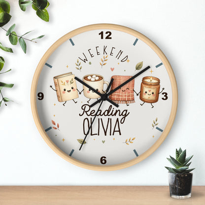 A round wall clock with a light wood frame and a white face, featuring a cute illustration of four books and a mug with faces, the text 'WEEKEND Reading OLIVIA', and black clock hands. The clock is shown hanging on a white wall above a wooden surface, with small green plants in the corners.