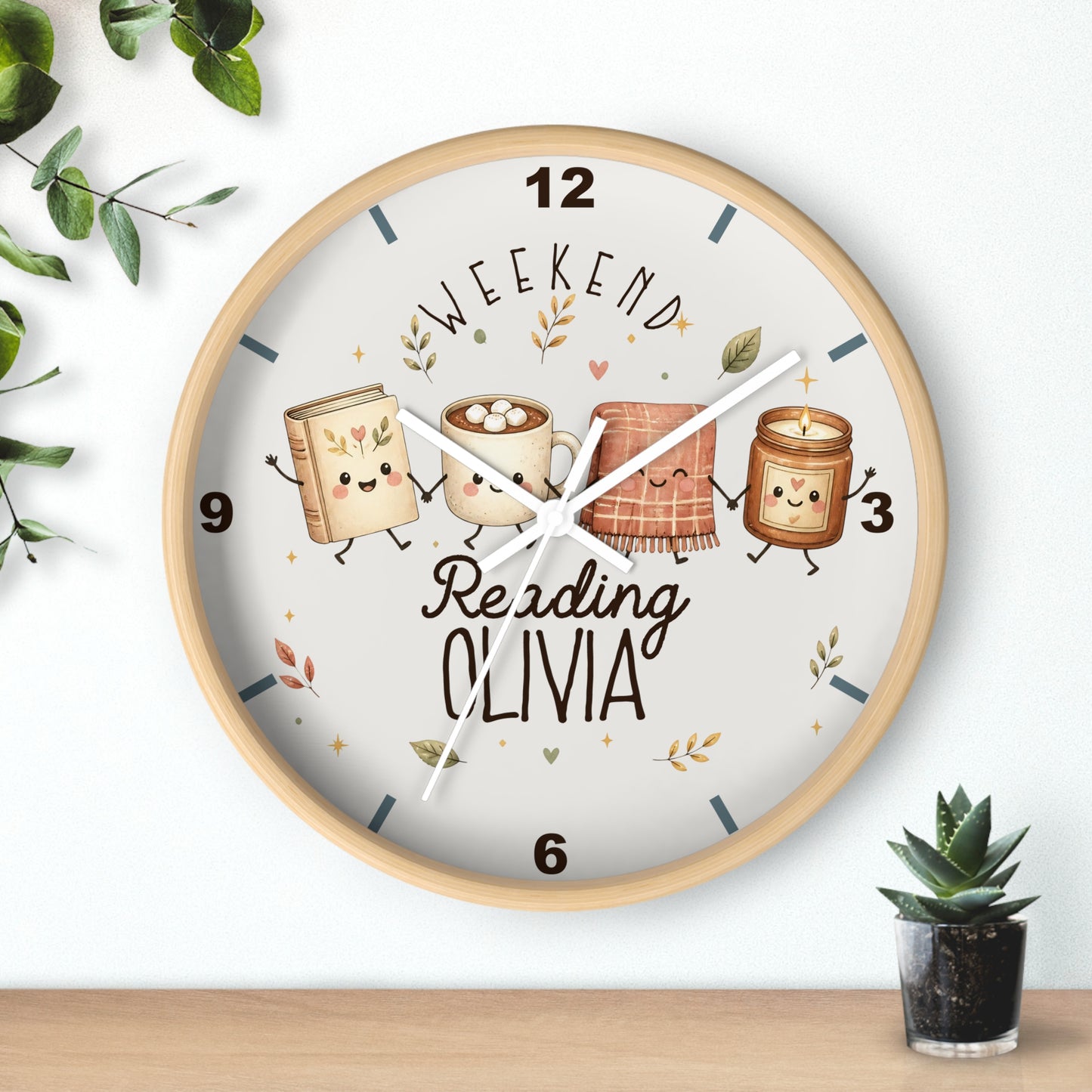 A round wooden wall clock with a light wood frame and a white dial, featuring cute illustrated books and a mug with the text 'WEEKEND Reading OLIVIA'. The clock is shown hanging on a white wall above a wooden shelf, with decorative green leaves and a small potted succulent.