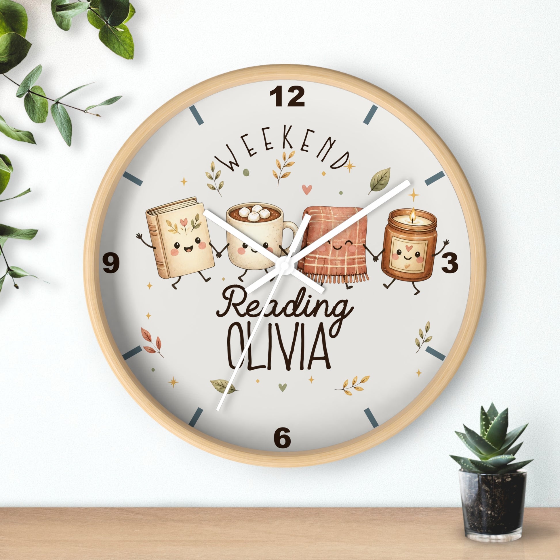 A round wooden wall clock with a light wood frame and a white dial, featuring cute illustrated books and a mug with the text 'WEEKEND Reading OLIVIA'. The clock is shown hanging on a white wall above a wooden shelf, with decorative green leaves and a small potted succulent.