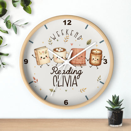 A round wooden wall clock with a light wood frame and a white dial, featuring cute illustrated books and a mug with the text 'WEEKEND Reading OLIVIA'. The clock is shown hanging on a white wall above a wooden shelf, with decorative green leaves and a small potted succulent.