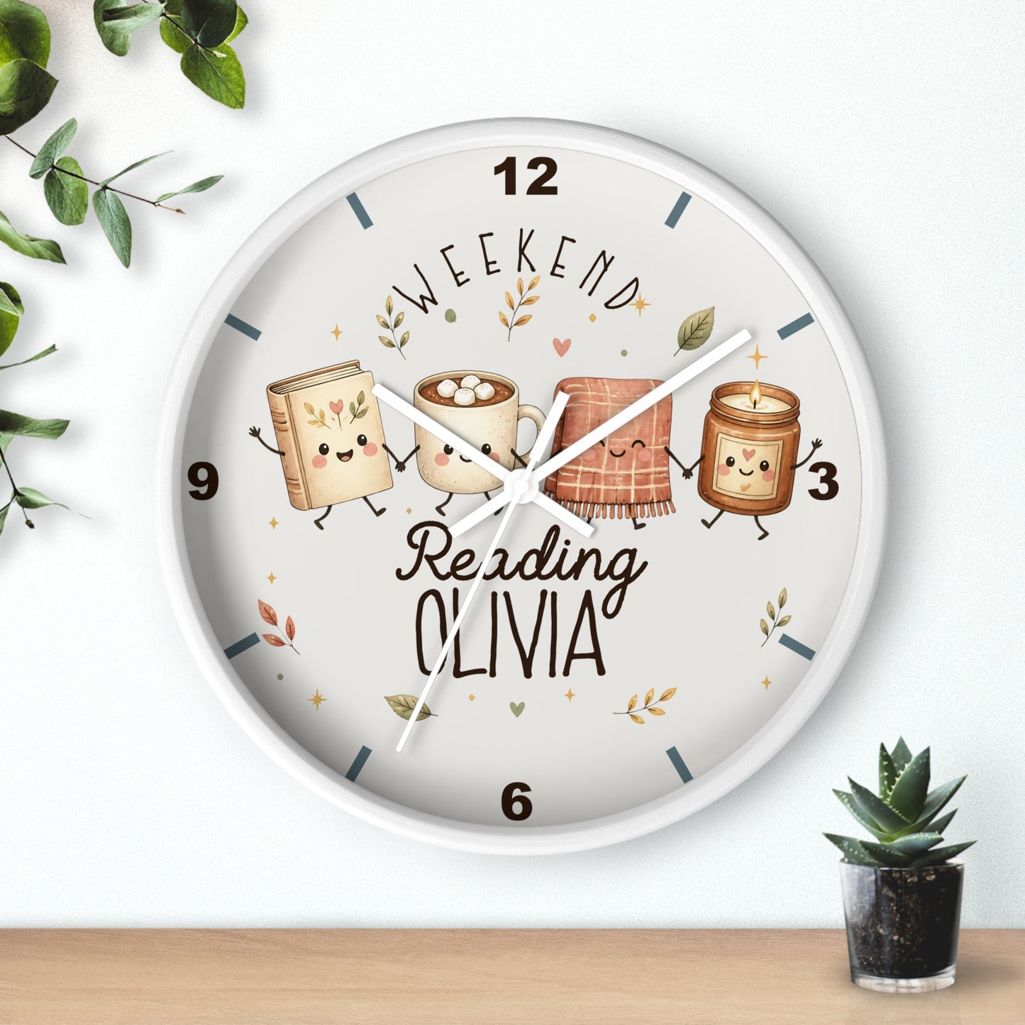 A round wall clock with a white frame and a whimsical printed face showing four marshmallow-themed books with faces, the words 'WEEKEND Reading OLIVIA,' and decorative leaves. The clock is mounted on a white wall above a light wood surface, with small green plants at the corners.