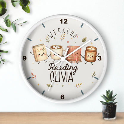 A round wall clock with a white frame and a whimsical printed face showing four marshmallow-themed books with faces, the words 'WEEKEND Reading OLIVIA,' and decorative leaves. The clock is mounted on a white wall above a light wood surface, with small green plants at the corners.