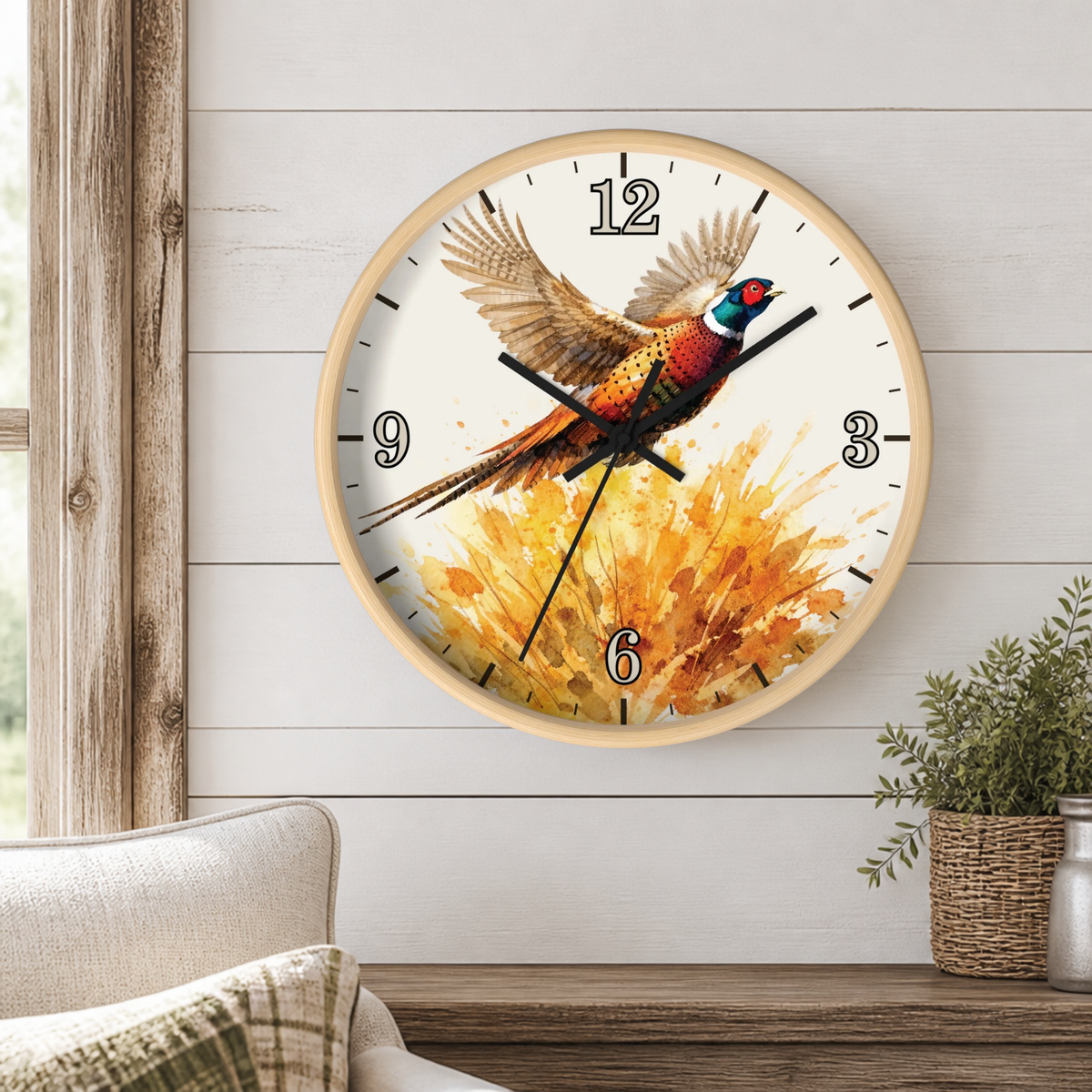A round wall clock with a light wood frame and a white face featuring a pheasant bird over a golden floral splash. The clock is mounted on a white shiplap wall above a wooden mantel with a small potted plant and part of a sofa visible.