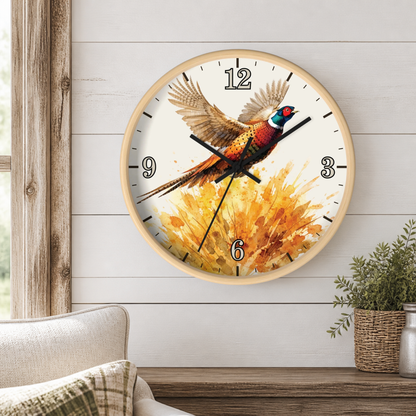 A round wall clock with a light wood frame and a white face featuring a pheasant bird over a golden floral splash. The clock is mounted on a white shiplap wall above a wooden mantel with a small potted plant and part of a sofa visible.