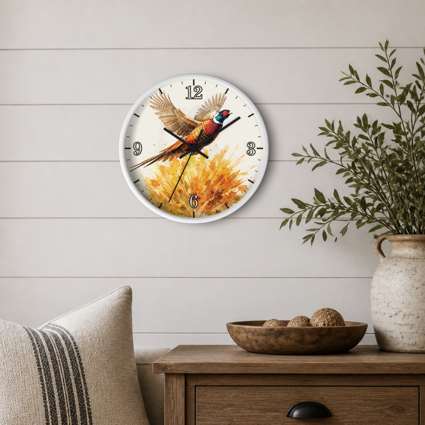 A round wall clock with a white frame hangs on a light wood shiplap wall above a wooden side table. The clock face features a colorful pheasant bird in flight over a golden-yellow floral background.