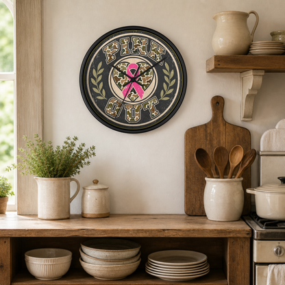 A round black-framed wall clock with a camouflage patterned face, featuring a pink ribbon and the words 'PINK OUT' in camouflage lettering, hanging on a beige wall in a rustic kitchen setting.