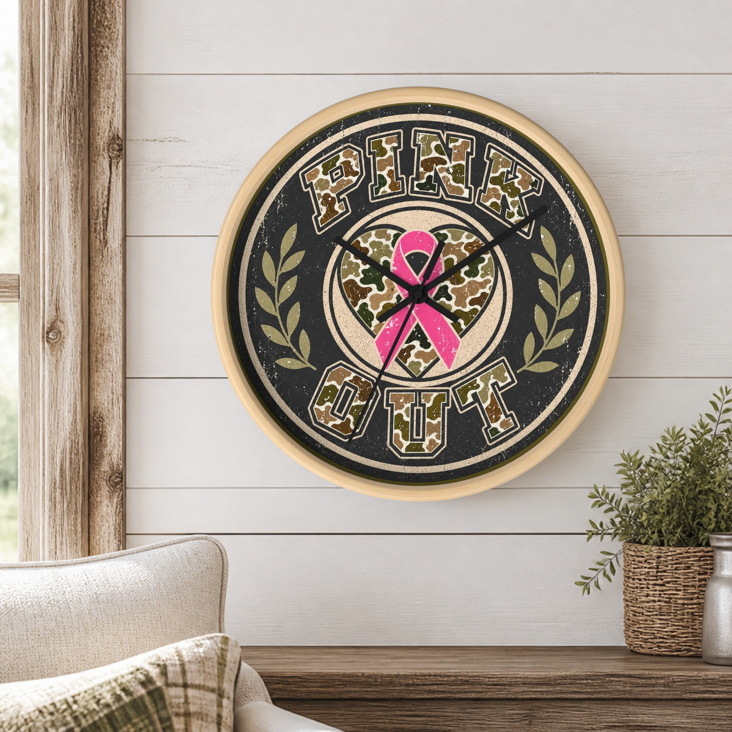 A round wall clock with a distressed design featuring the words 'PINK OUT' and a pink ribbon over a camouflage pattern, mounted on a white shiplap wall in a styled room setting.