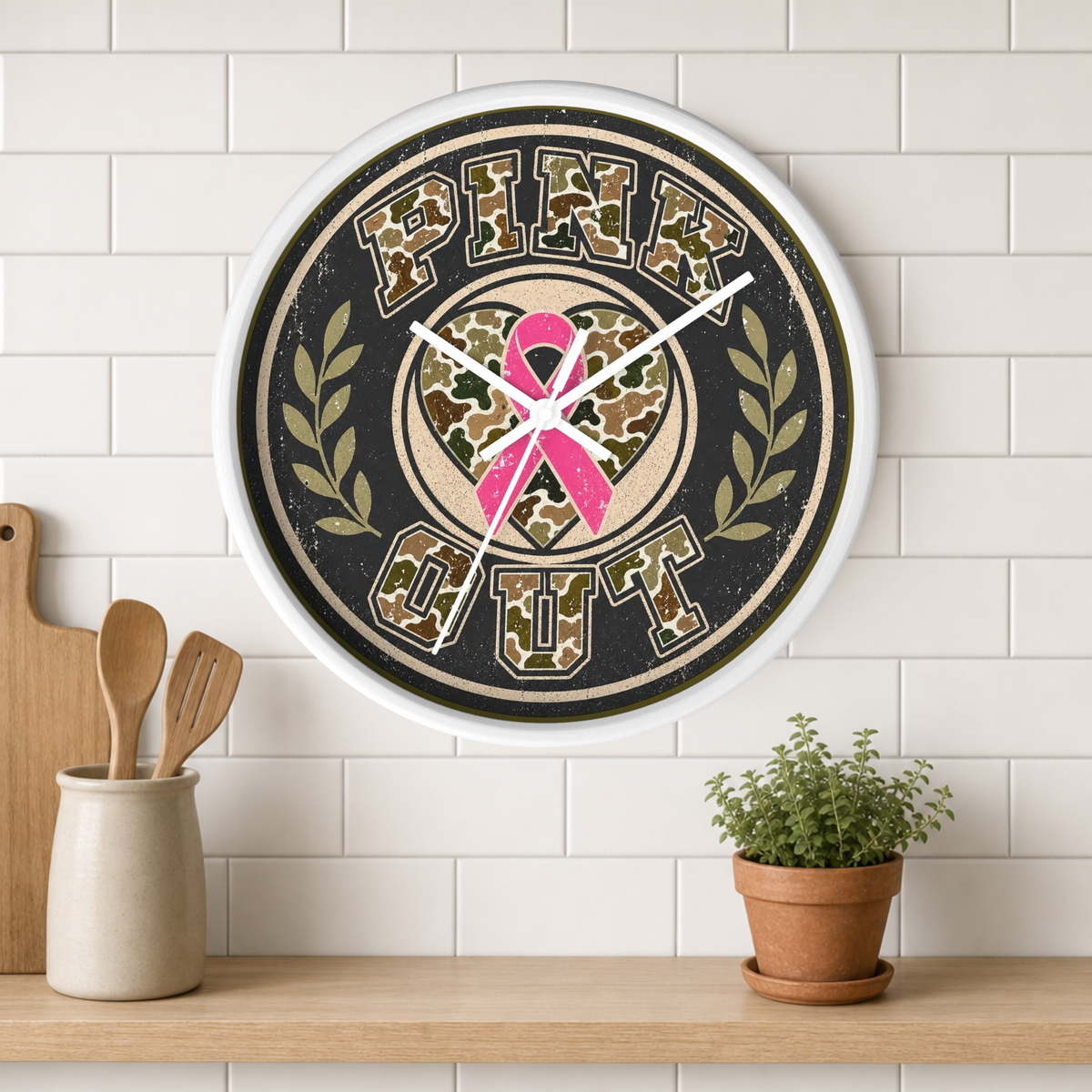 A round wall clock with a distressed black and beige design, featuring the words 'PINK OUT' in block letters around a camouflage heart with a pink ribbon in the center. The clock is mounted on a white subway tile wall above a wooden shelf with kitchen utensils and a potted plant.