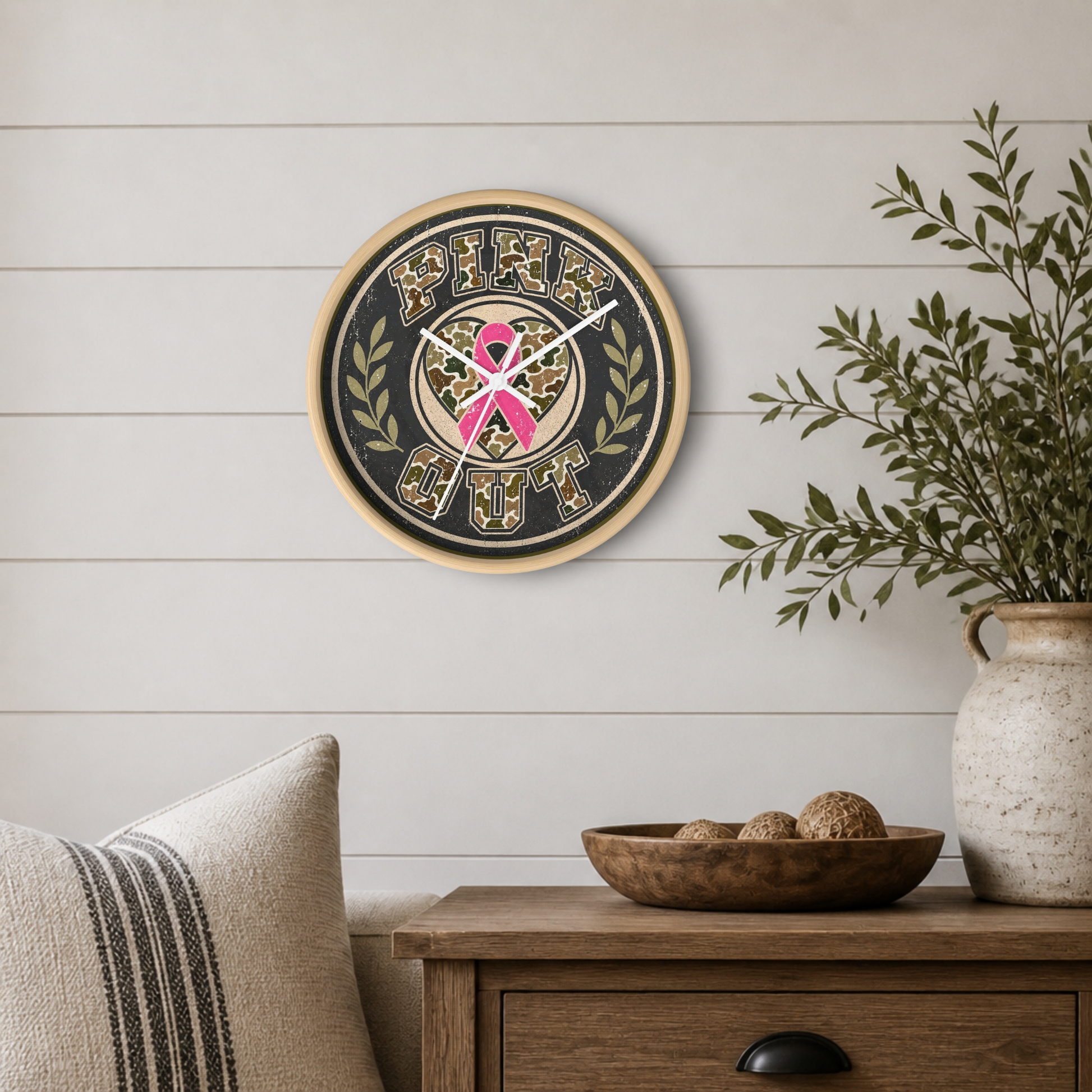 Round wall clock with a pink ribbon and camouflage pattern, mounted on a white shiplap wall above a wooden side table with a plant and decorative items.