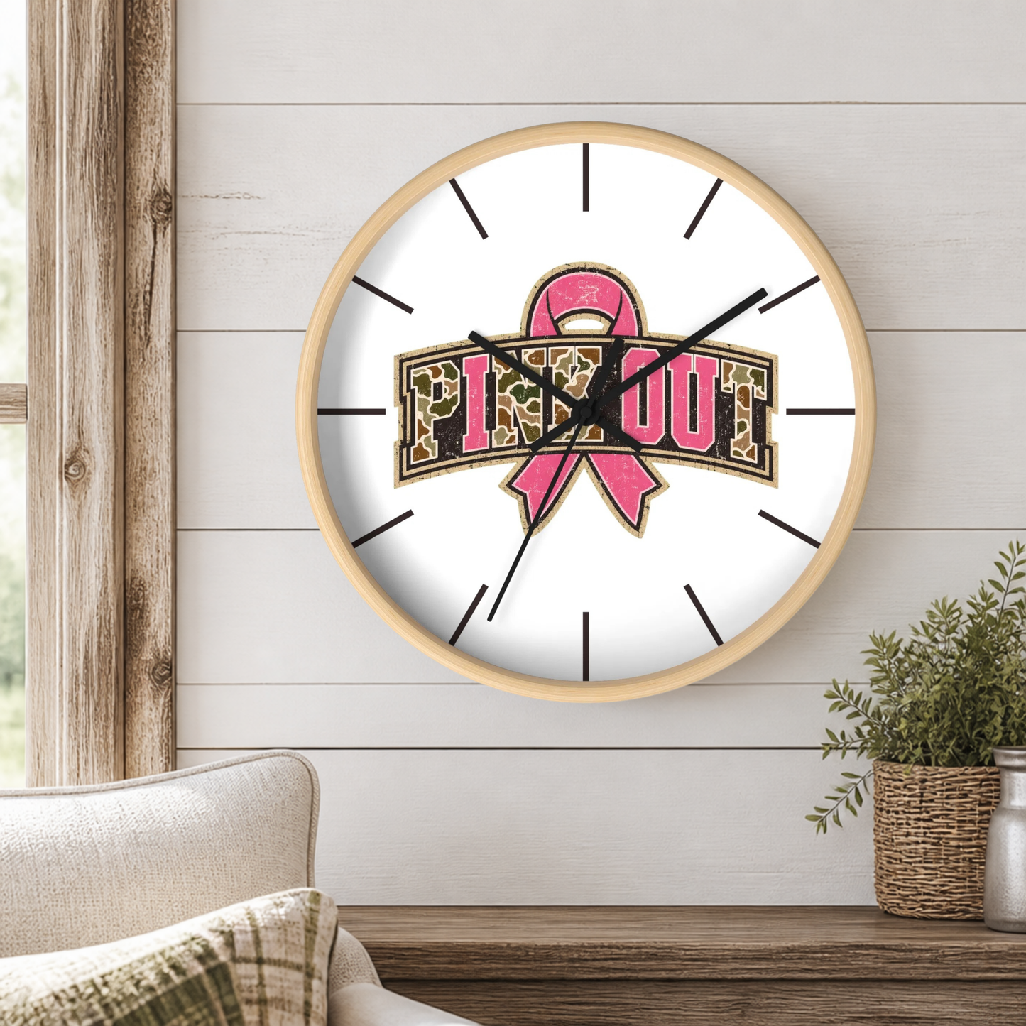 A round wall clock with a light wood frame hangs on a white shiplap wall above a wooden table. The clock face is white with black hands and stick-style hour markers, featuring a pink awareness ribbon and the words 'Pink OUT' in leopard-print lettering.