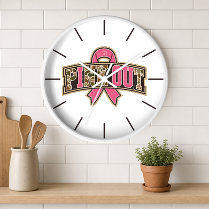 A round white wall clock with a pink ribbon and leopard-print 'PINK OUT' graphic, mounted on a white subway tile wall above a wooden shelf with a small potted plant and kitchen utensils.