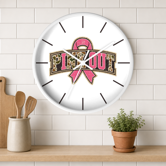 A round white wall clock with a pink ribbon and leopard-print 'PINK OUT' graphic, mounted on a white subway tile wall above a wooden shelf with a small potted plant and kitchen utensils.