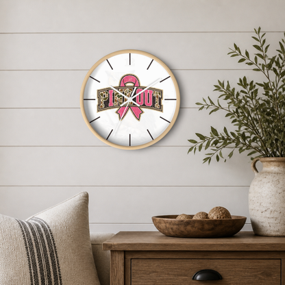 A round wall clock with a light wood frame hangs on a white shiplap wall above a wooden side table. The clock face is white with black stick-style hour markers and hands, featuring a pink ribbon and a gold-colored graphic across the center.