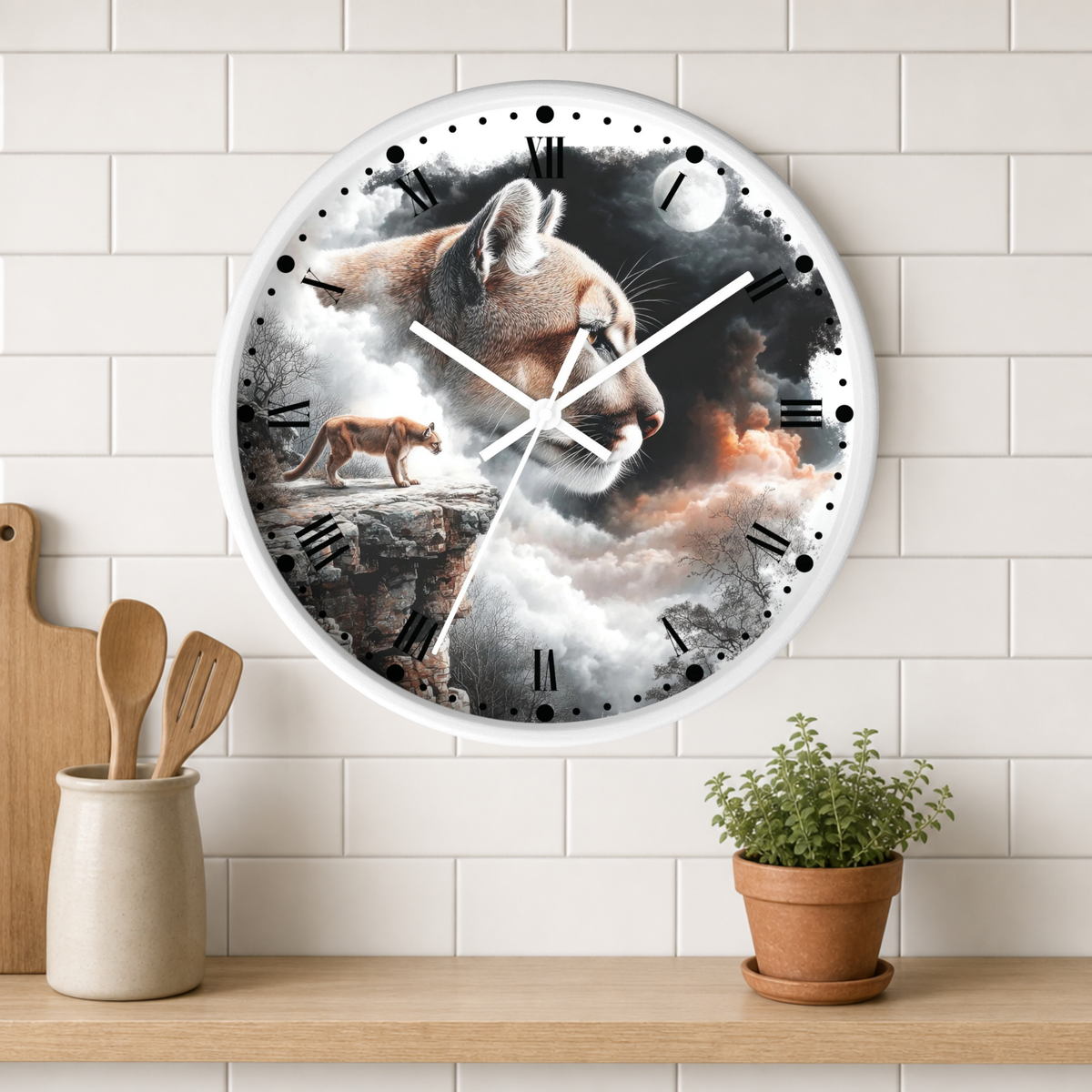A round wall clock with a white frame hangs on a white subway-tile wall above a wooden shelf. The clock face features a dramatic illustration of a mountain lion in profile, with a smaller lion cub standing on a rock in the background, set against a cloudy sky with a moon.