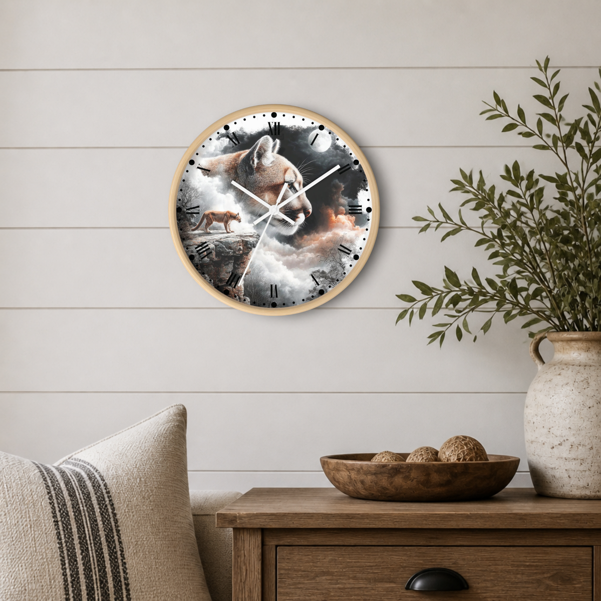 A round wall clock with a light wood frame hangs on a white shiplap wall above a wooden side table. The clock face features a dramatic painting of a wolf howling at a moon, with a deer on the left and a warm orange glow near the center.
