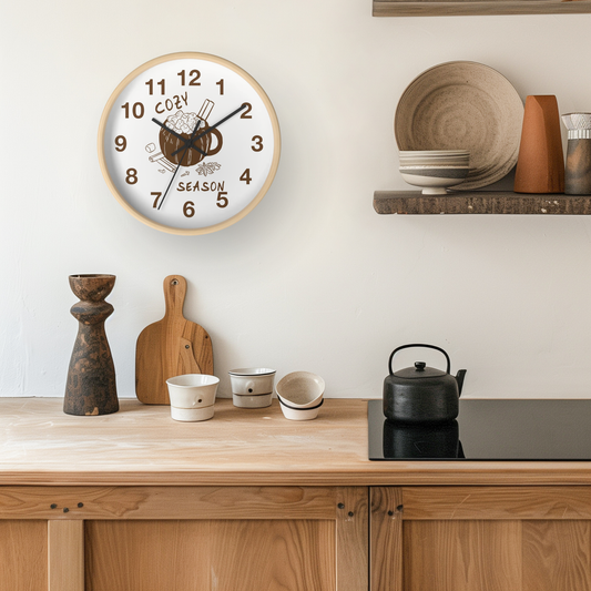 A round wall clock with a light wood frame hangs on a white wall above a wooden kitchen counter. The clock face is white with black Arabic numerals and a central illustration of a mug with steam and the words 'COZY SEASON'.